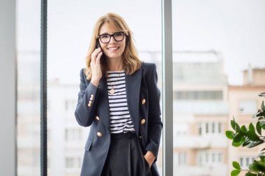 Akıllı telefon kullanan olgun bir kadın. Ofiste dikilirken kendine güveni tam bir iş kadını arıyor. Gözlük ve spor ceket giyen çekici bir kadın. Boşluğu kopyala.