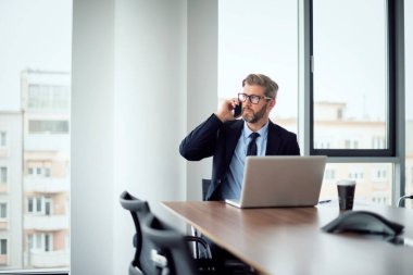 Yetişkin bir iş adamı, dizüstü bilgisayarla masada oturuyor ve cep telefonuyla konuşuyor. Ofiste çalışan iş adamı..