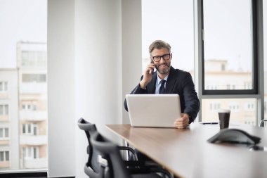 Yetişkin bir iş adamı, dizüstü bilgisayarla masada oturuyor ve cep telefonuyla konuşuyor. Ofiste çalışan iş adamı..