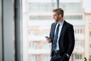 Ofiste dururken akıllı telefon ve kulaklık kullanan gülümseyen iş adamının portresi. Gözlüklü ve takım elbiseli profesyonel bir adam.. 