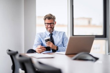 Yetişkin bir iş adamı, dizüstü bilgisayarla masada oturuyor ve cep telefonuyla konuşuyor. İş adamı ofiste çalışırken video görüşmesi yapıyor.