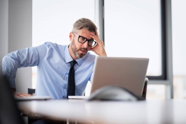 Ofiste çalışıp dizüstü bilgisayar kullanan orta yaşlı bir işadamı. Profesyonel adam endişeli görünüyor..