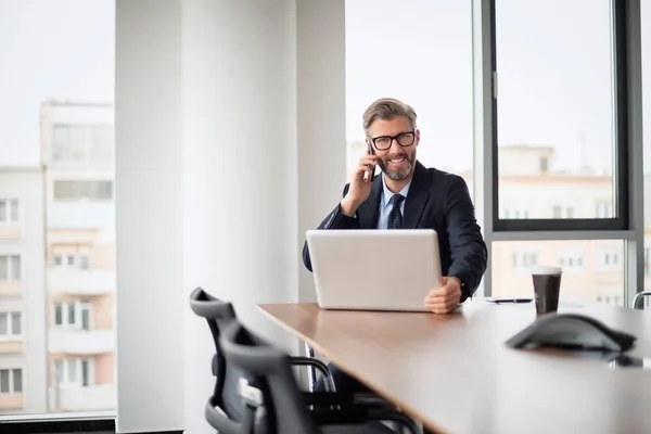 ミッド大人のビジネスの専門家は ラップトップでテーブルに座って 携帯電話で話しています 事務所で働くビジネスマン — ストック写真