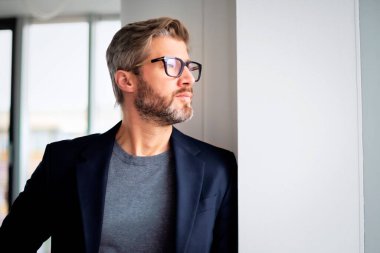Close-up of thinking businessman wearing a sweater with a suit while standing at the office and looking out the window