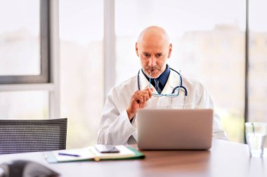 Shot of a mature doctor sitting alone in his clinic and looking contemplative while using his laptop. 