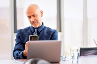 Shot of a thinking businessman using mobile phone and laptop while sitting at office desk and working. Middle aged professional man wearing shirt and tie. 