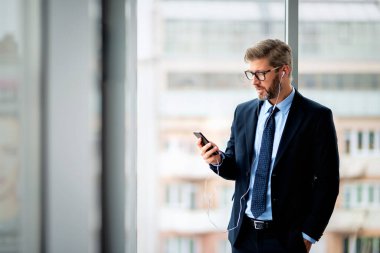 Confident businessman using smartphone and earphones while standing at the office. Professional man wearing suit and tie.