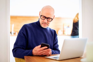 Thinking man text messaging and using laptop while working from home. Confident male sitting in the kitchen and text messaging. Home office.