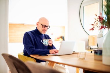Confident middle aged man using laptop and having video call while working from home. Confident male drinking tea while sitting at table at the kitchen. Home office.