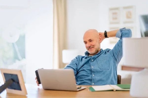 Confident middle aged man using laptop and having video call while working from home. Happy male sitting at desk and wearing shirt. Home office.