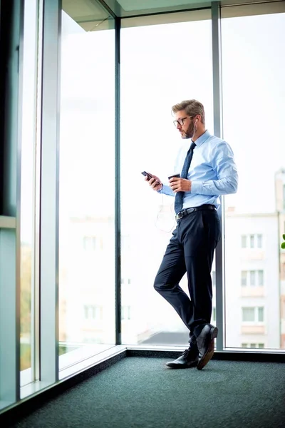 Full length of businessman using smartphone and earphones while standing at the office. Professional businessman text messaging at the office and drinking coffee.