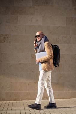 Mature businessman using mobile phone in the city. Man in casual clothes walking outdoors by a building. Full length shot. 