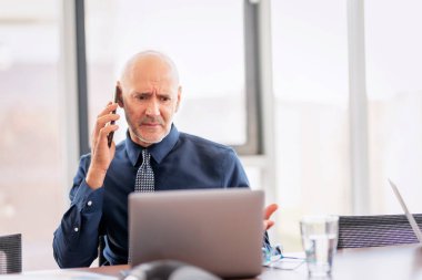 Shot of middle aged businessman looking stressed while using a laptop and smartphone in a modern office and talking with somebody.