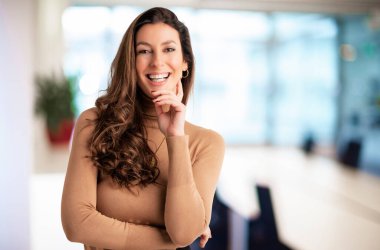 Close-up of an attractive middle aged female cheerful smiling and looking at camera. Brunette haired businesswoman wearing sweater while standing at the office. Copy space. 