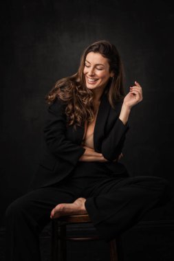 Full length of an attractive mid aged woman with toothy smile wearing black blazer and pants and sitting against at isolated dark background. Copy space. 