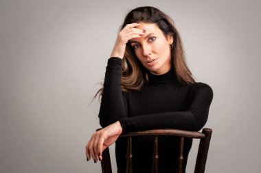 Close-up of an attractive middle aged woman wearing turtleneck sweater while sitting at isolated dark background. Hand on forehead. Copy space. Studio shot.