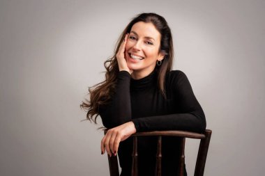 Close-up of an attractive middle aged woman with toothy smile wearing turtleneck sweater while sitting at isolated grey background. Copy space. Studio shot.