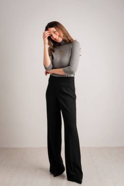 Full length of an attractive mid aged woman smiling and looking at camera. Brunette haired female wearing turtleneck sweater and pants while standing at isolated background. Copy space. 