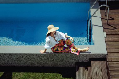 From above shot of unrecognizable woman wearing straw hat and white shirt while relaxing at the swimming pool. Shot from above. 