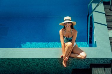 Full length of a happy woman wearing swimsuit and straw hat while relaxing at the poolside. 