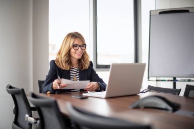 Smiling mid aged businesswoman sitting at the office and having video conference. Blond haired professional female wearing blazer and glasses and holding a document in her hand. 