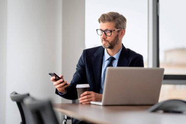 Thinking business professional sitting at table with laptop and mobile phone and text messaging. Professional man wearing suit while working at the office.