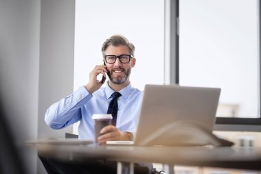 Mid aged businessman sitting at office desk and having a business call. Confident professional man drinking coffee and using notebook.