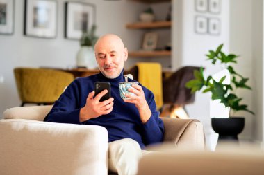 Evdeki koltukta dinlenirken cep telefonu ve mesaj kullanan orta yaşlı bir adamın portresi. Balıkçı yaka kazak giyen ve çay içen mutlu bir erkek..