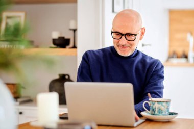 Orta yaşlı bir adam dizüstü bilgisayar kullanıyor ve evde çalışırken video görüşmesi yapıyor. Mutfaktaki masada kendinden emin bir erkek oturuyor. Merkez Ofis.
