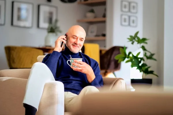 中年男子在家中坐在扶手椅上放松时使用手机和打电话的肖像 快乐的男性穿着高领毛衣喝茶 — 图库照片