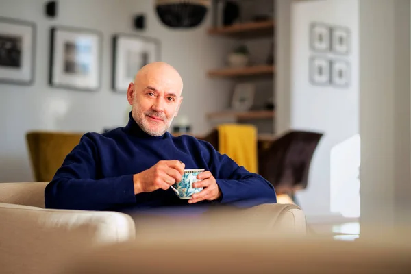 自宅のアームチェアでリラックスしてお茶を飲む中年男性の肖像画 幸せな男性身に着けていますTurtleeckセーターと笑顔 — ストック写真