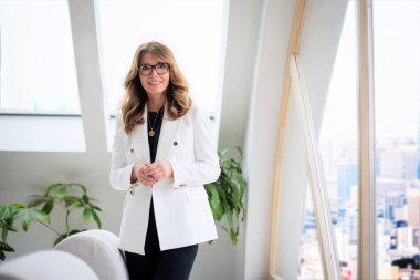 Çekici, orta yaşlı bir iş kadınının fotoğrafı. Kameraya bakıyor ve pencerenin önünde gülümsüyor.. 