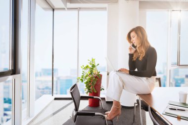 Ofiste oturan çekici bir iş kadını modern bir ofiste otururken cep telefonu ve dizüstü bilgisayar kullanıyor..