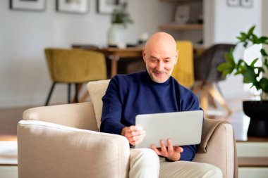 Modern bir evde koltukta oturan orta yaşlı bir adam iş için dizüstü bilgisayar kullanıyor. Merkez Ofis.