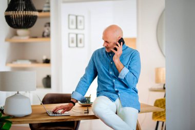 Mutlu orta yaşlı bir adam evde çalışırken telefon edip laptop kullanıyor. Oturma odasında masasında duran kendinden emin bir erkek. Merkez Ofis.