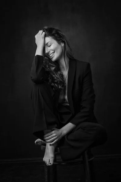 黒のブレザーとパンツを背景に隔離された暗い背景に座っ歯の笑顔で魅力的な中年女性のフル長さ スペースのコピー — ストック写真