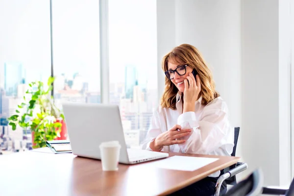 中年女商人坐在一个现代化的办公室里 有一个商务电话 工作时使用笔记本电脑的自信的职业女性 — 图库照片