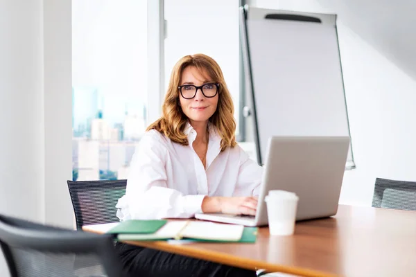 微笑着坐在现代化的办公室里 用笔记本电脑工作的中年女商人 穿着眼镜和白衬衫的金发职业女性 — 图库照片
