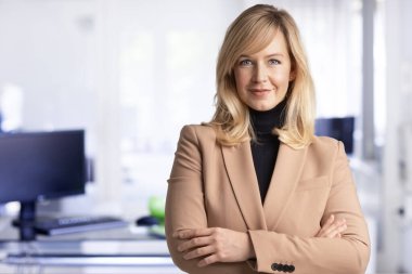 Orta yaşlı iş kadınının gülümseyen ve kameraya bakan yakın çekimi. Ofiste dikilirken spor ceket giyen sarışın bir kadın. Boşluğu kopyala. 