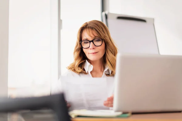 在现代化的办公室工作时 中年职业女性穿着衬衫 使用笔记本电脑 迷人的女商人坐在写字台前 — 图库照片