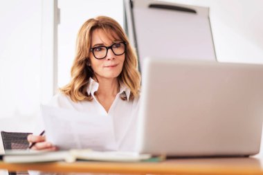 Modern bir ofiste çalışırken orta yaşlı, profesyonel bir kadın tişört giyiyor ve laptop kullanıyor. Sarı saçlı iş kadını. Gözlük takıyor.. 