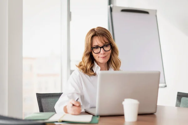 中年职业女性使用笔记本电脑 在现代化的办公室举行视频会议 金发女商人 头戴眼镜 身穿白衬衫 还在工作 — 图库照片