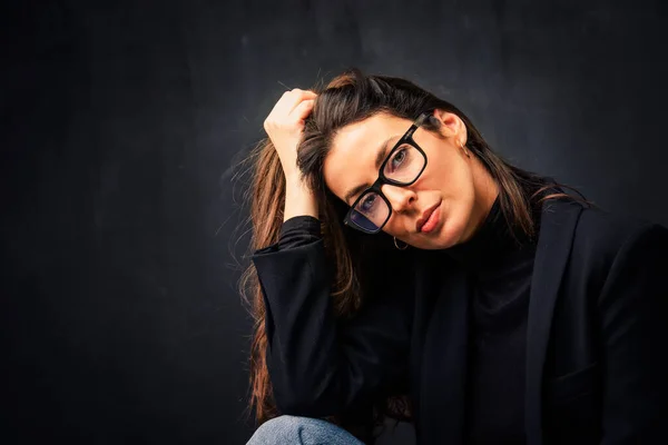 stock image Headshot of an attractive middle aged woman wearing eyeglasses and black blazer while posing at isolated dark background. Copy space. Studio shot. Hand on head.