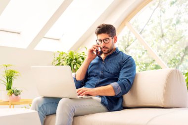 Evden çalışırken akıllı telefon ve dizüstü bilgisayar kullanan bir adam. Kendine güveni tam bir erkek koltukta oturuyor ve telefonla konuşuyor. Merkez Ofis.
