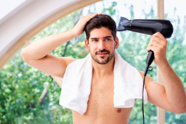 Saçları ıslak yakışıklı adam banyo aynasının önünde duruyor ve saçını kurutuyor.. 