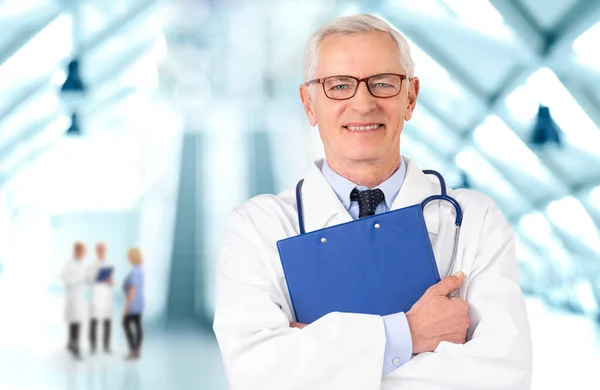 病院の廊下に立ちながら 研究室のコートと聴診器を身に着けているシニア男性医師と彼の手にクリップボードを保持 — ストック写真