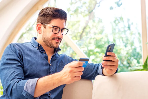 집에서 소파에 온라인 쇼핑에 휴대폰 카드를 사용하는 잘생긴 남자의 — 스톡 사진