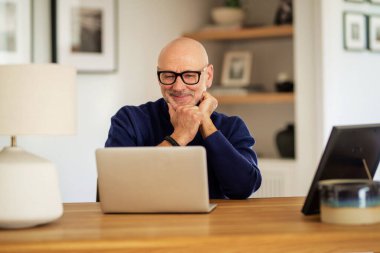 Thinking middle aged man using laptop and having video call while working from home. Confident male sitting at table at the kitchen. Home office.