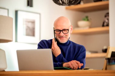 Confident middle aged man making a call and using laptop while working from home. Confident male sitting in the kitchen and having a call. Home office.