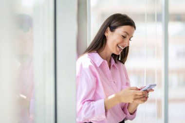 Esmer, orta yaşlı bir iş kadını modern bir ofiste duruyor ve mesajlaşıyor. Tişört giyen ve neşeli gülümseyen kendine güvenen profesyonel bir kadın.. 